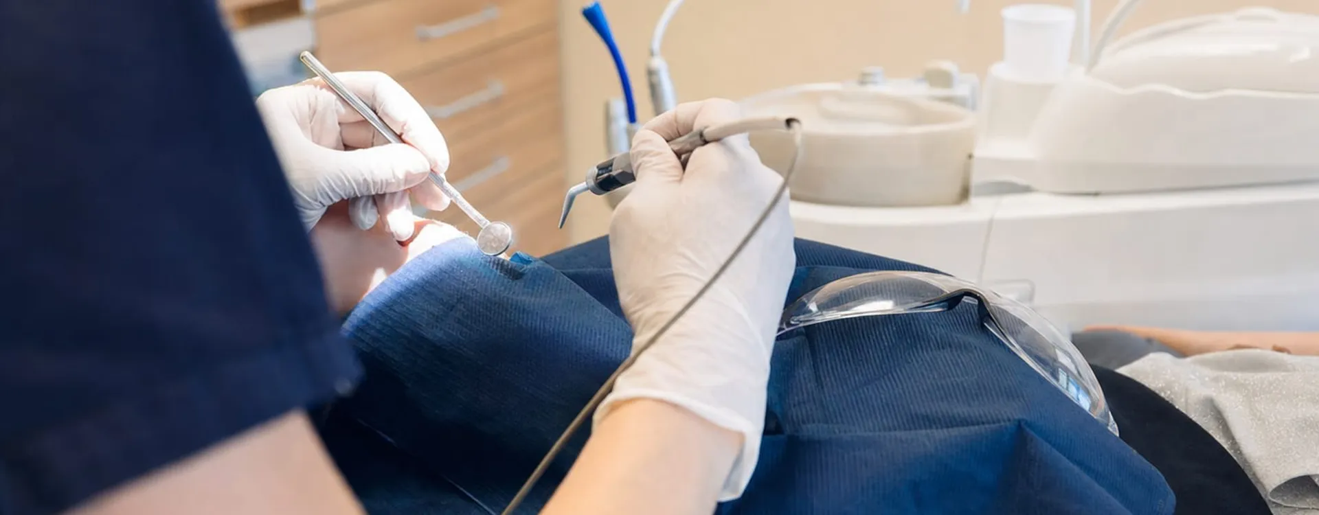 Zahnmedizinische Fachkraft in dunkelblauer Kleidung und weißen Handschuhen behandelt einen Patienten mit Spiegel und Instrument im Behandlungszimmer.